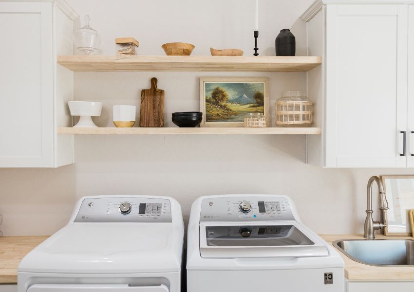 A new and bright white washer and dryer are pictured underneath two wooden shelving units, and between white wooden cabinets.